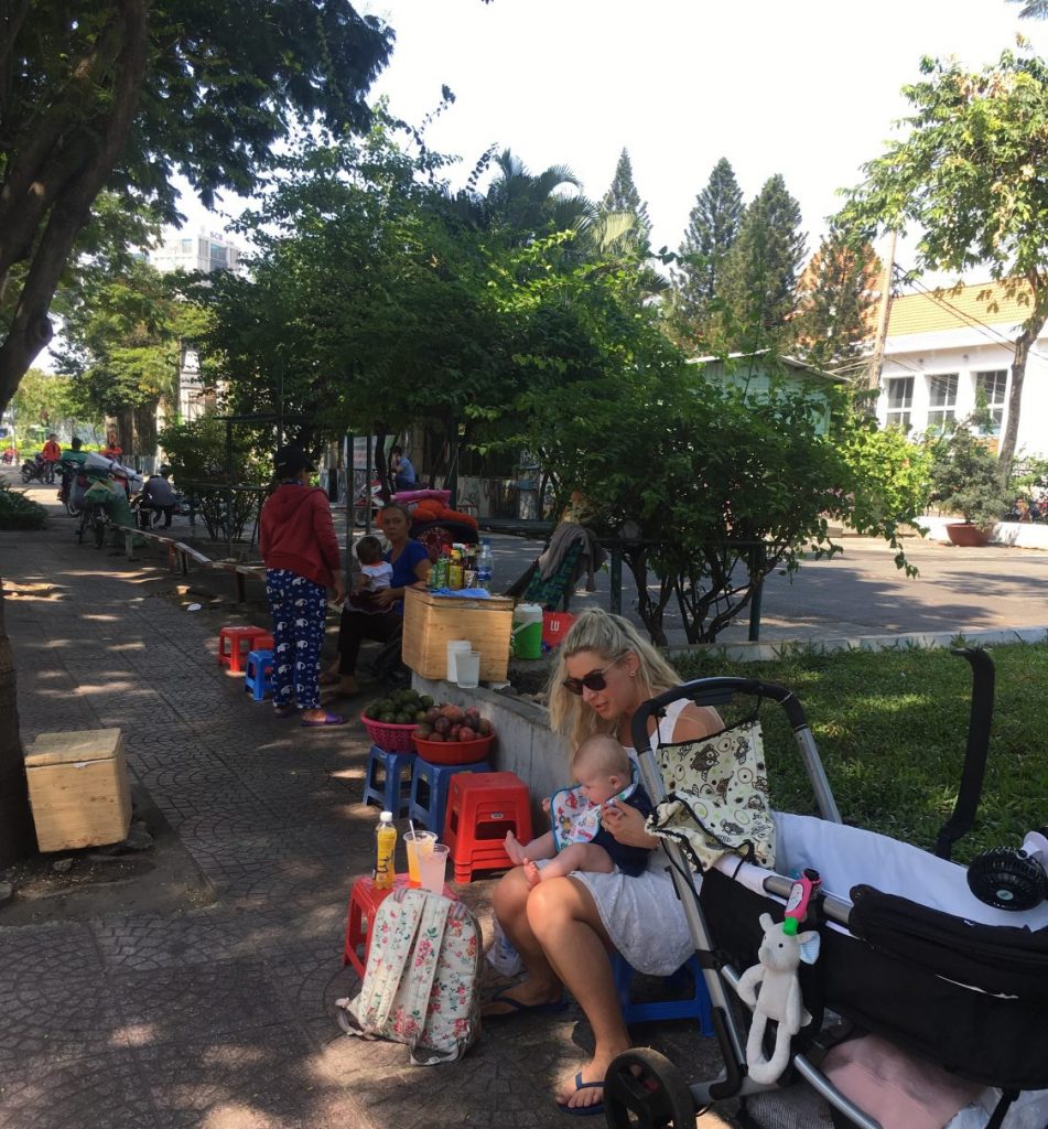 A mother holding her newborn baby taking a break from the heat in Ho Chi Minh City in Vietnam. She is sat on a roadside cafe, on a little blue childs stool, while the female street vendor makes fresh fruit to attract other customers