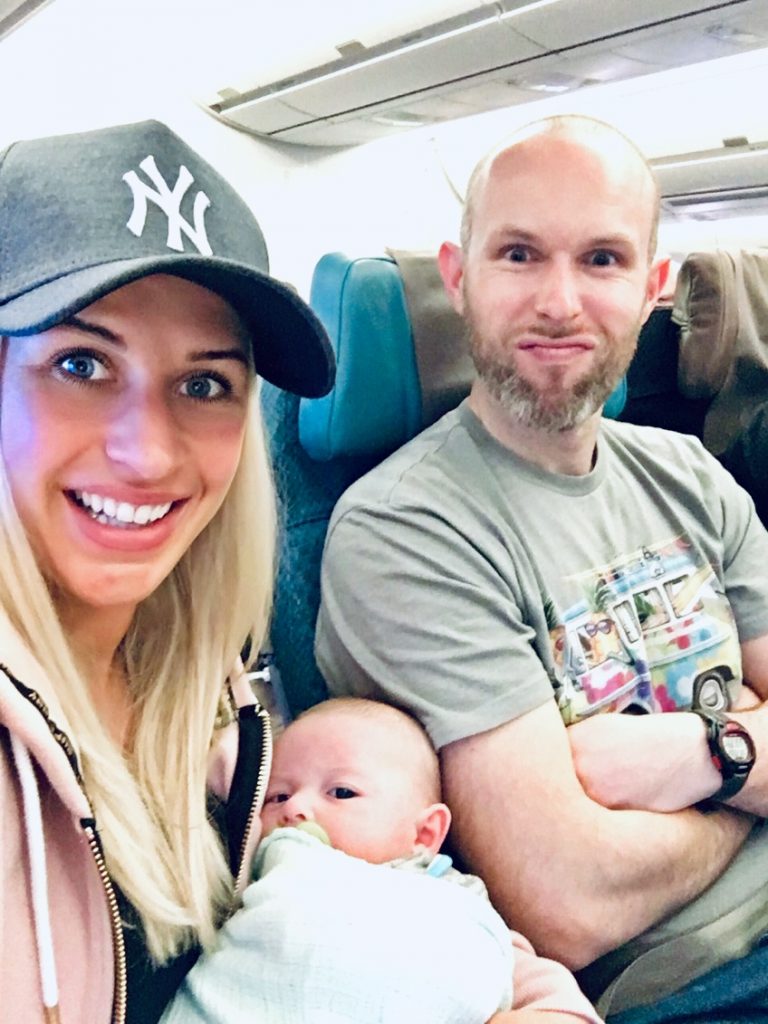 Two parents sat side by side on a plane holding a young baby in their arms. Both are smiling. 