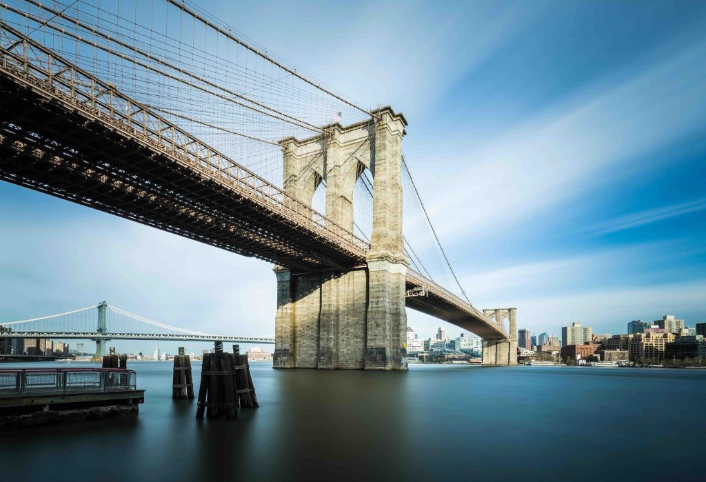 A panoramic view of the famous Brooklyn Bridge during the day. Contrasting blue of the sky clashing with the hard brown of the construction brick. 
