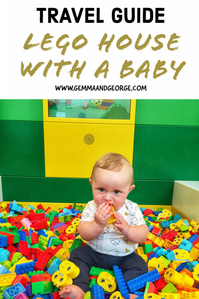 A young baby boy is sat in a pool of lego, looking at the camera eating a Lego Duplo brick. 