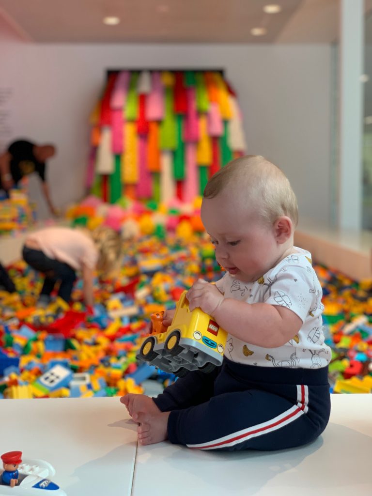A young male baby is sat upright and playing with a yellow Lego truck. Behind him is a rainbow waterfall made entirely of lego bricks. He is smiling whilst playing at Lego House in Billund, Denmark.