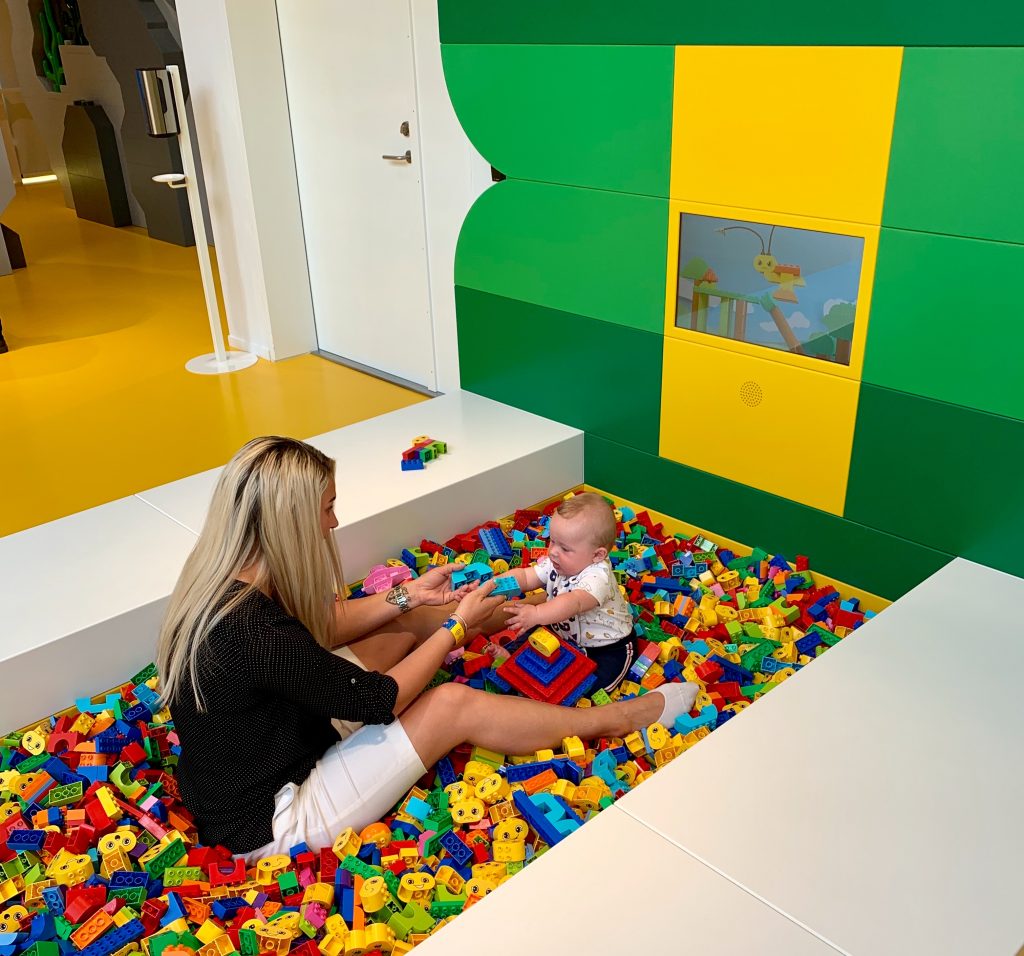 A mother and son play in a big pool of multi-coloured Lego Duplo bricks. The young son is handing her bricks as they sit opposite each other. 