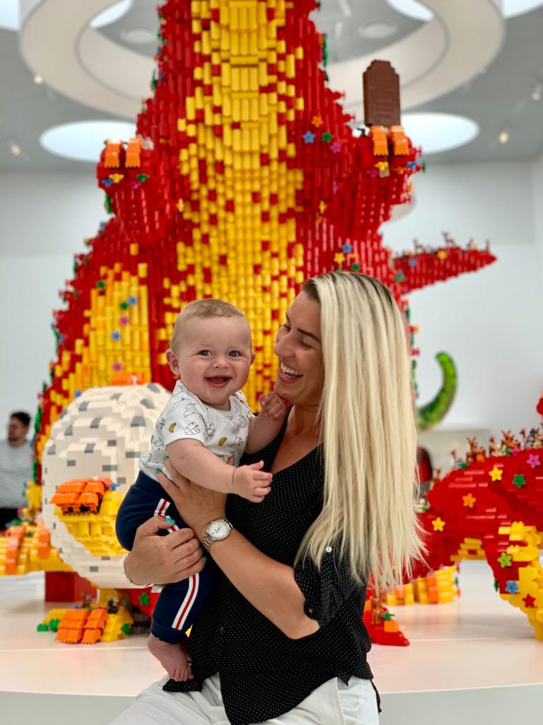 A mother holds her son in her arms and he smiles at the camera, looking very happy. The pair are stood infront of a large size replica of a dinosaur, made entirely from Lego Bricks. On display at Lego House in Billund, Denmark. 
