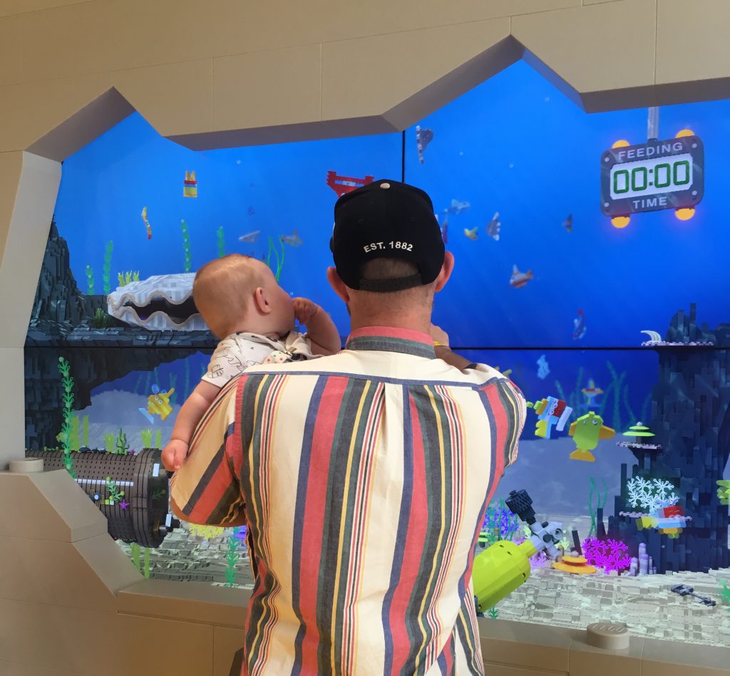 A father and son, looking at an interactive fish tank in Lego House in Billund. The father holds the young baby in his arms and points out fish that swim by.